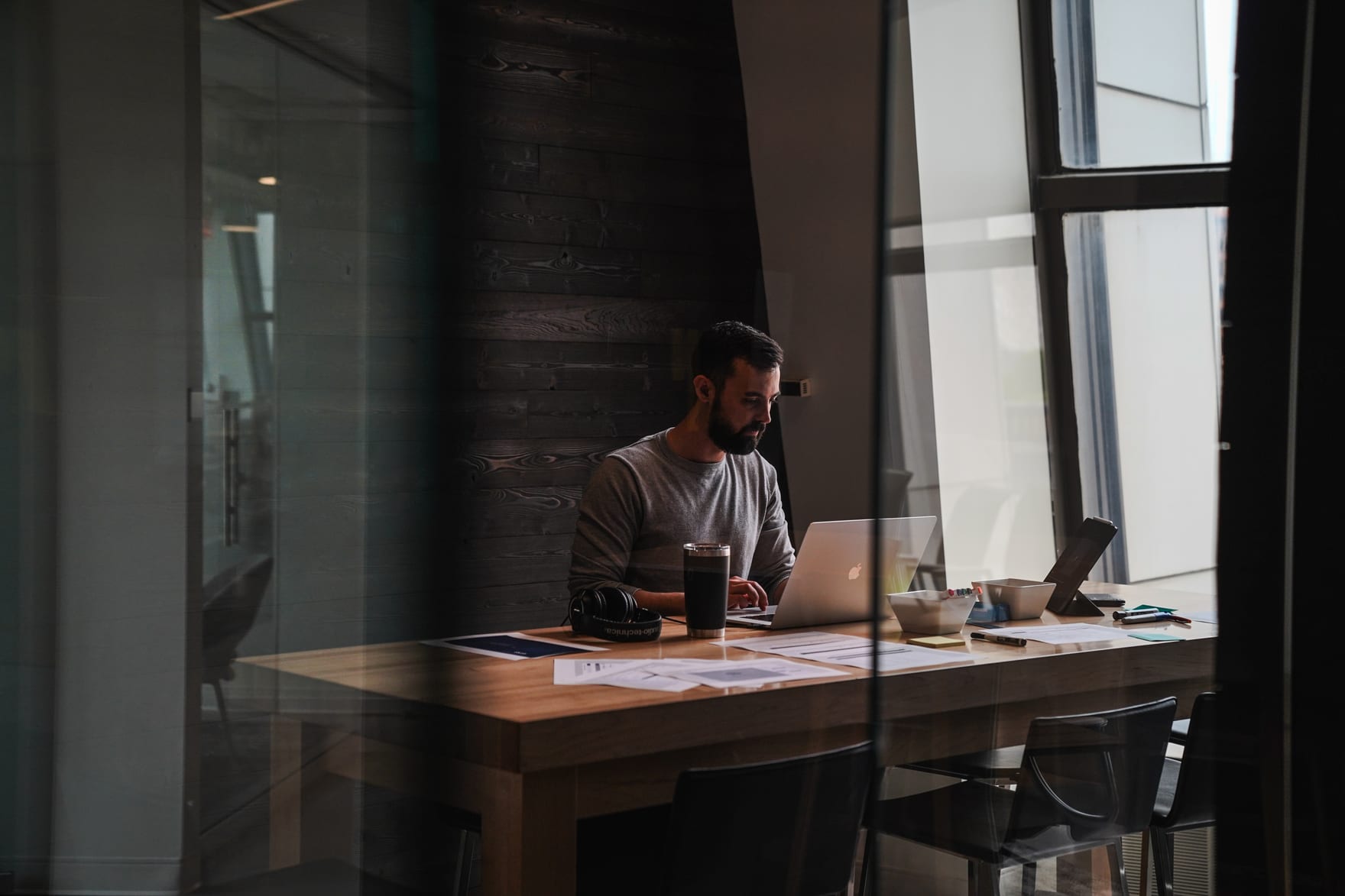David working at a laptop at a long wooden table, seen through a glass partition