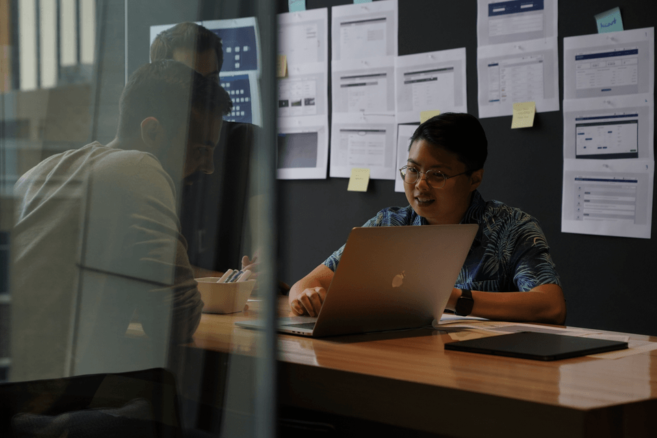Two people working at a laptop in a meeting room, with printed wireframes and sticky notes pinned to the wall behind them