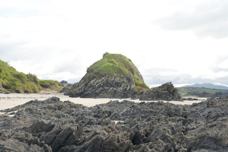 Grass-topped rock formation rising from a beach, jagged rocks in foreground