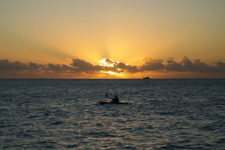 A kayaker paddling open water as the sun breaks through clouds at sunrise