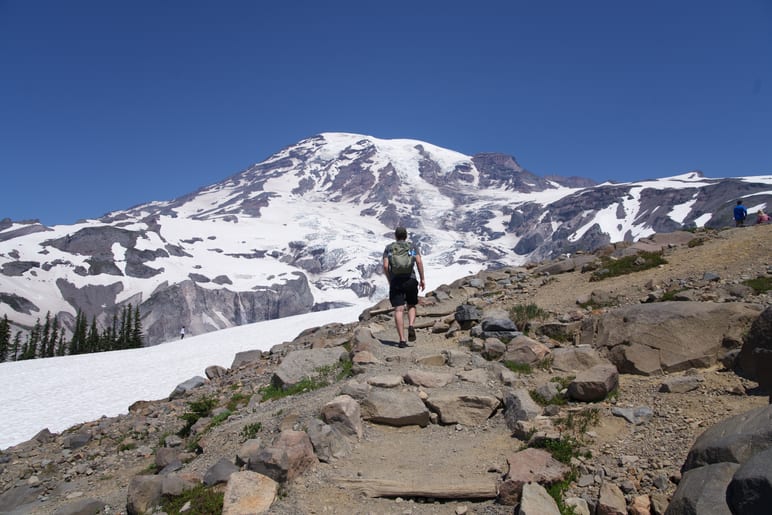A hiker walking a rocky trail toward a snow covered volcano