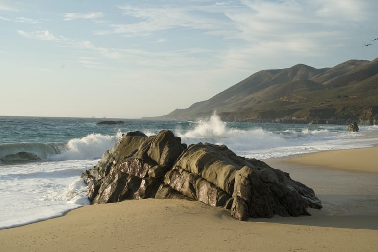 Waves crashing around a large dark rock on a sandy beach, hills behind