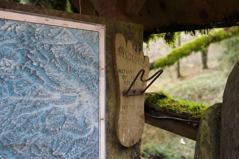 A carved wooden sign and trail map hang under a rustic shelter roof