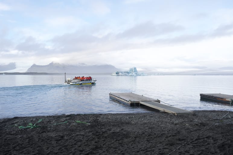 A small tour boat crosses calm water near floating icebergs under cloudy sky