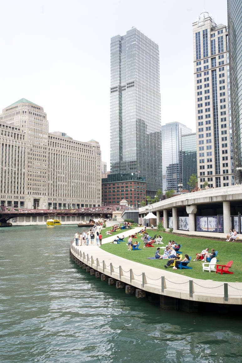 People relaxing on a riverside green lawn framed by tall glass skyscrapers