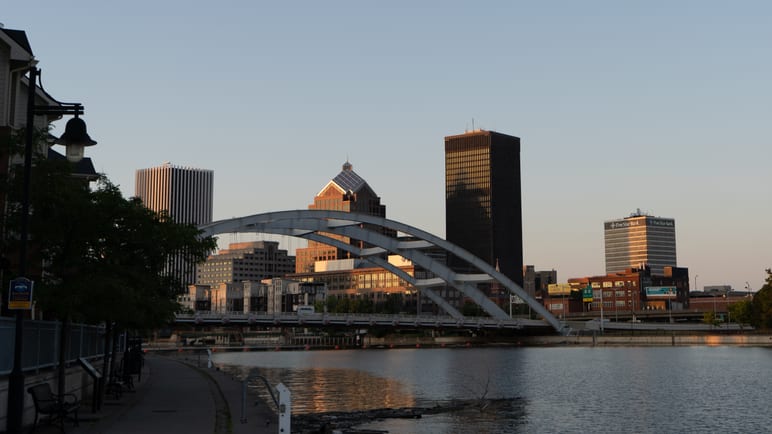 A city skyline and an arched pedestrian bridge reflected in a river at dusk