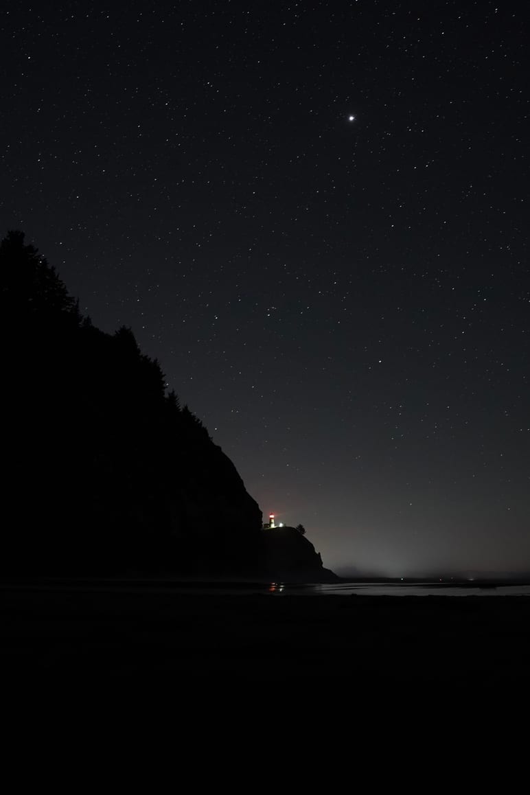 Starry night sky above a dark cliff with a small light at its base