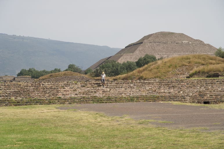 Person walking along a low stone wall with a stepped pyramid beyond, hazy hills behind