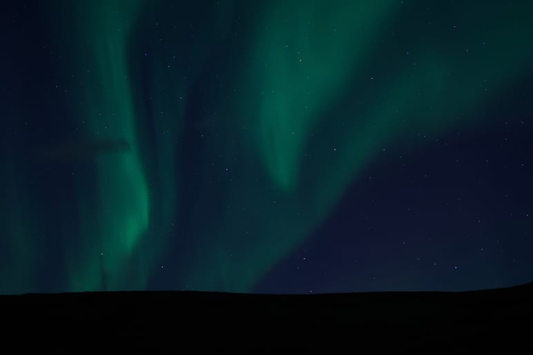 Green aurora ribbons swirl across a starry night sky above a dark ridge