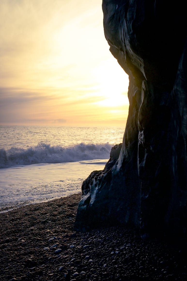 Sunset glows through a dark rock formation onto a pebbled beach with crashing waves