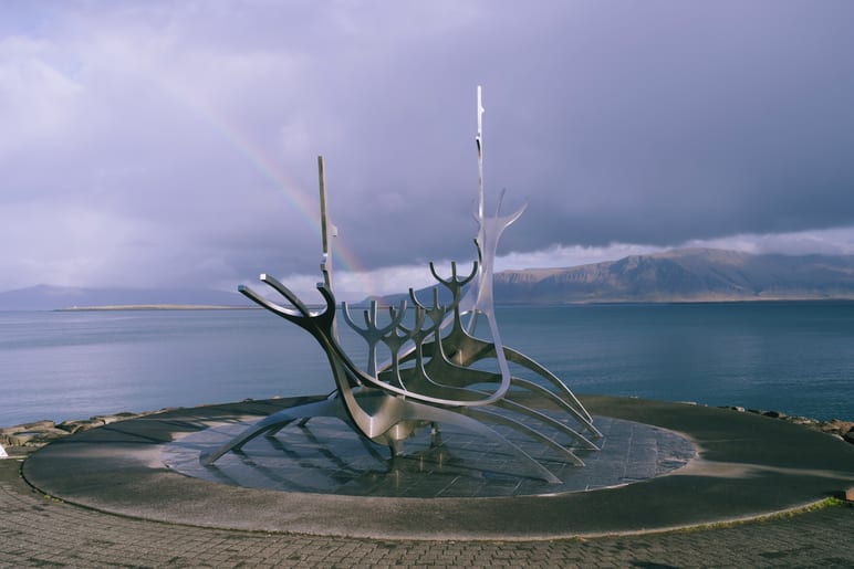 Metal ship sculpture on a waterfront plaza under a rainbow and clouds