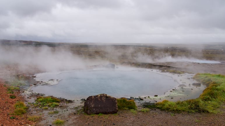 Steam rises from a blue geothermal pool surrounded by misty ground