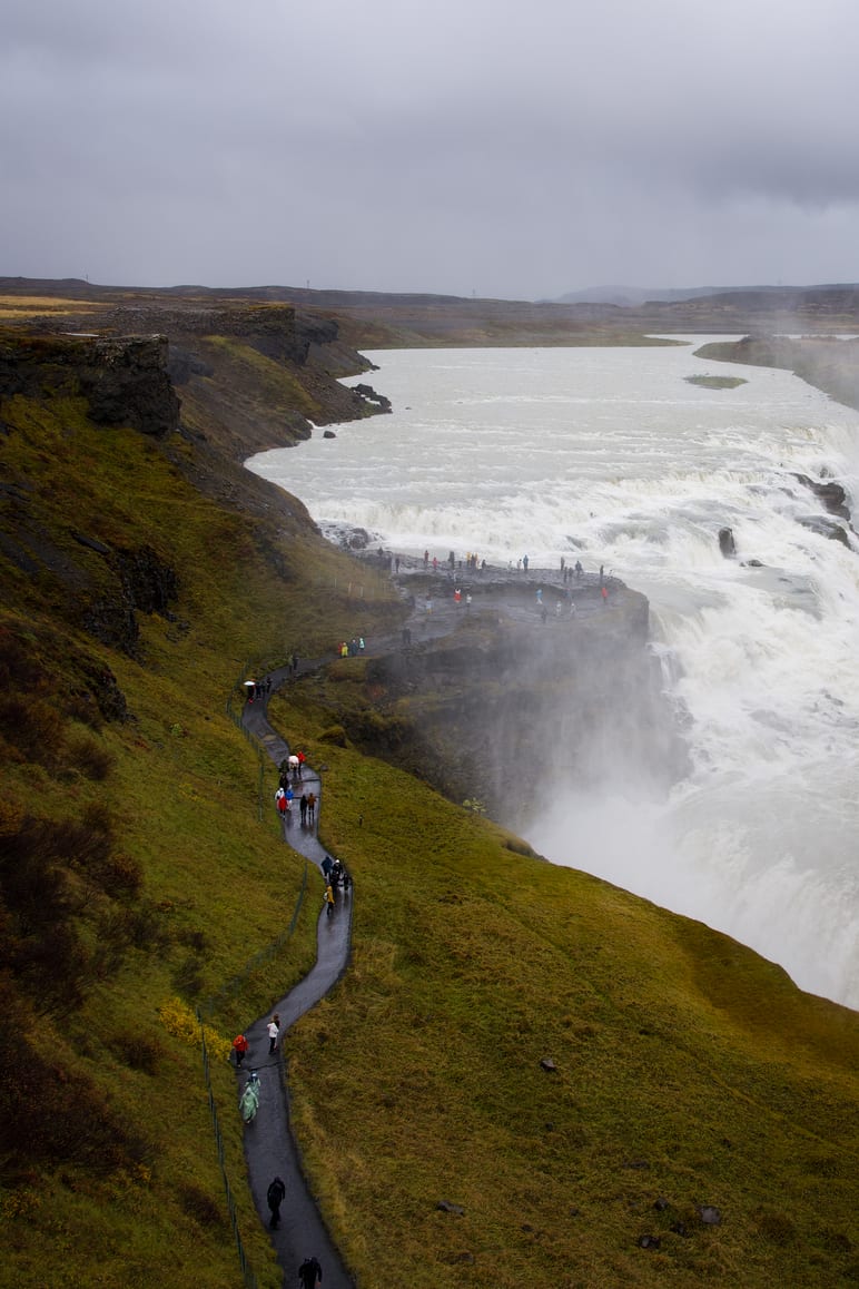 Visitors walk a path along a cliff overlooking a wide waterfall