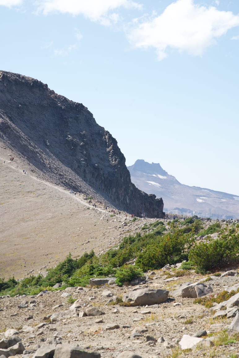 Hikers on a narrow trail along a steep rocky ridge with a peak beyond