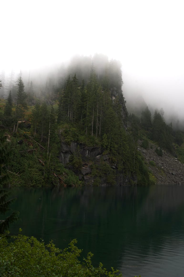 A forested cliff wrapped in fog above a still lake