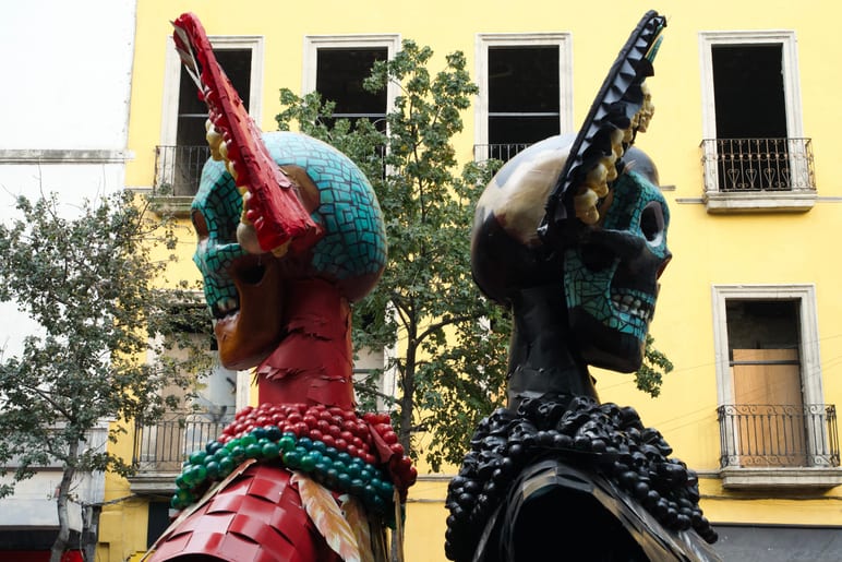 Two large decorated skull effigies in colorful headdresses before a yellow building