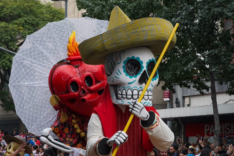 Towering skeleton parade puppets in a sombrero and red heart costume, crowd below